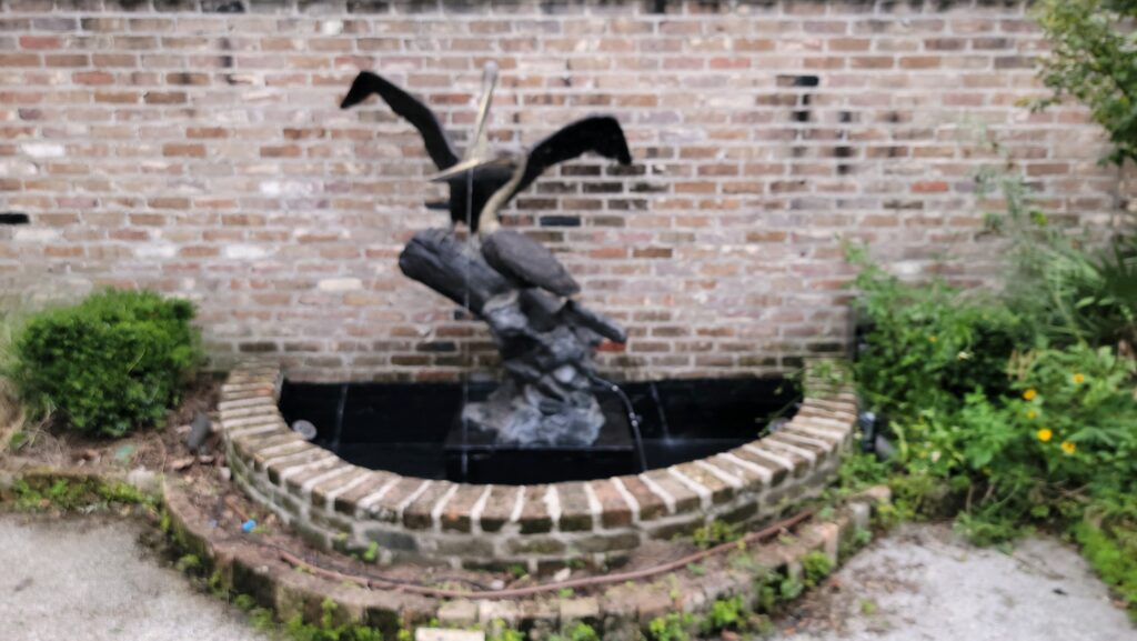 A fountain with a brick wall and plants in the background.